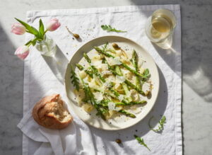 Asparagus carpaccio raw with parmesan shavings and lemon oil on a cream-colored plate, Pinot Grigio white wine glass, northern Italian marble setting