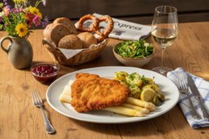 White asparagus with veal escalope and parsley potatoes on an oval porcelain plate, Bavarian Sunday light, white wine glass slightly misted over