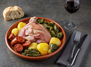 Lacón con grelos on clay plate, pork pink and juicy, grelos dark green, chorizo red, potatoes yellow-white, red wine glass with mencía dark red, dark gray granite, Atlantic winter light, oblique bird's eye view
