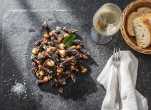 Percebes al natural on dark wooden board from above, barnacles gleaming with creamy white meat, sea salt next to them, white wine glass heavily misted, dark gray granite, diffuse Atlantic light, overhead